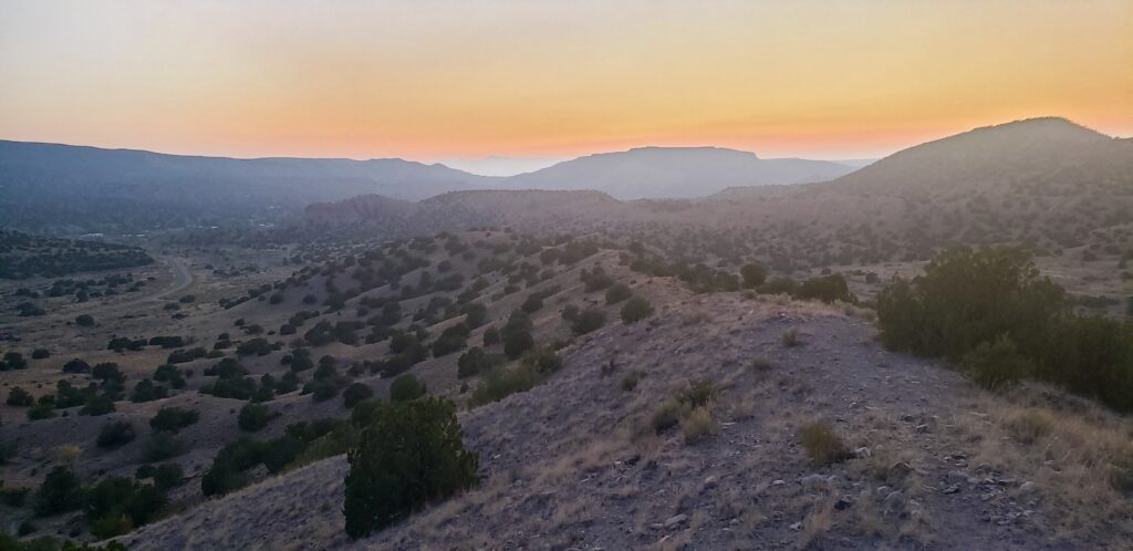 A rocky ridge in the foreground drops into a wide valley of juniper-dotted hills, layered mesas fading blue into an orange and pink sunset sky.