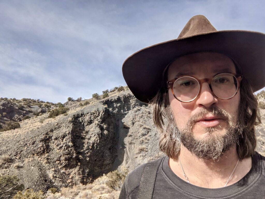 Close view of a bearded man in a wide-brimmed brown felt hat and round tortoiseshell glasses, looking into the camera. Behind him a dark grey rock face cuts across a pale sage-dotted slope under a thin-clouded blue sky.