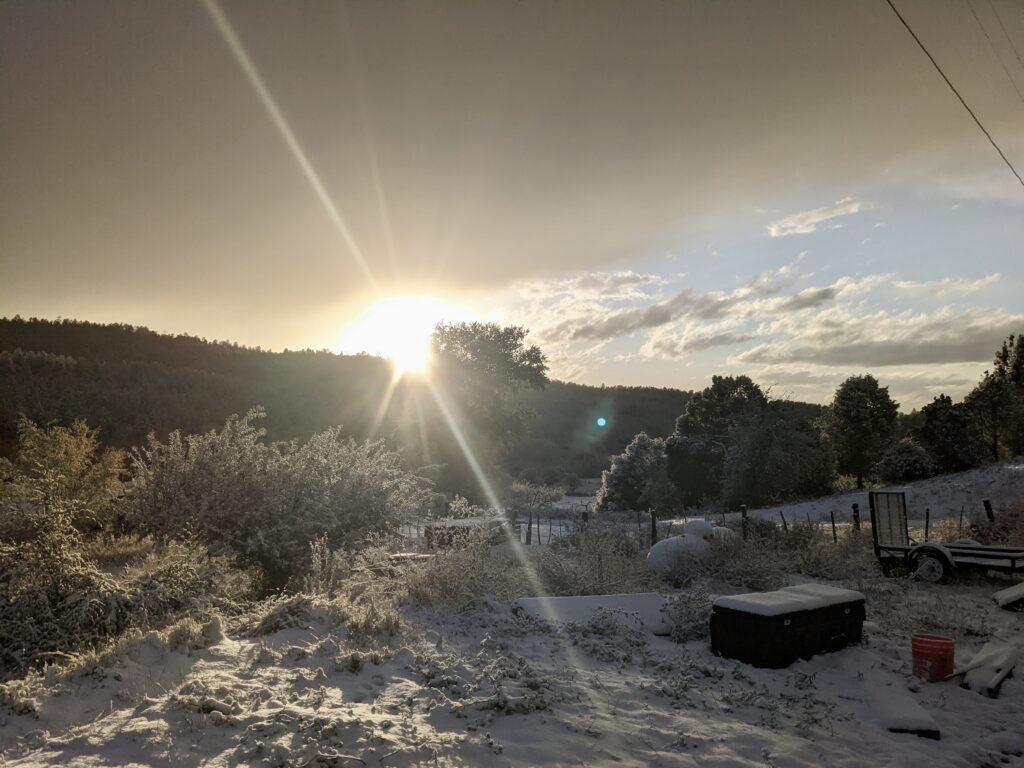 Sun breaking over a forested ridge, its rays flaring across a snow-covered yard with frost-coated shrubs, a wire fence, a propane tank, and a small trailer.