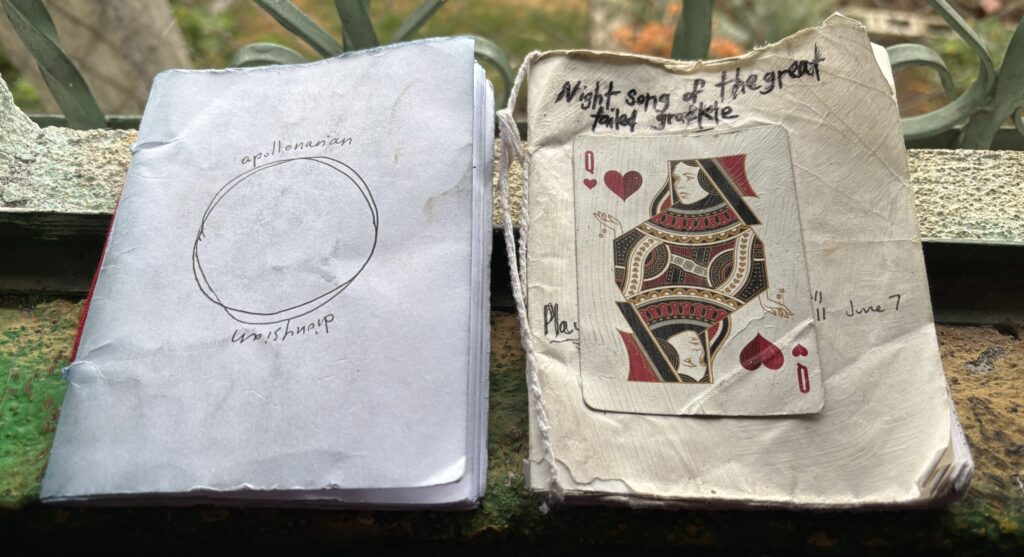 Two hand-bound notebooks of folded paper on a windowsill. One cover has a hand-drawn circle labeled in pen; the other is worn soft, titled in marker, with a Queen of Hearts playing card glued to the front.